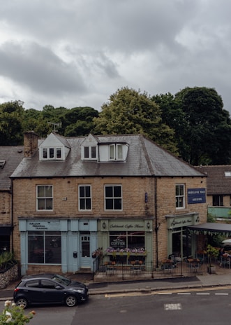 A two-story building made of stone bricks, featuring small businesses on the ground floor including a personal training center, a coffee shop with outdoor seating, and an interior design store. The sky is overcast with clouds, and there is a parked car on the street in front of the building. Lush green trees are visible in the background.