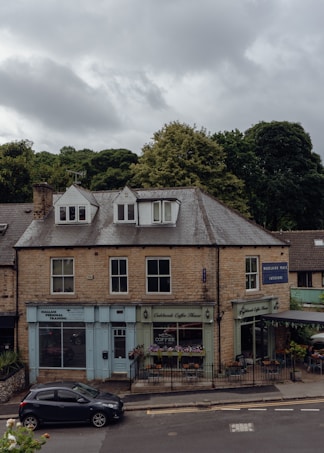 A two-story building made of stone bricks, featuring small businesses on the ground floor including a personal training center, a coffee shop with outdoor seating, and an interior design store. The sky is overcast with clouds, and there is a parked car on the street in front of the building. Lush green trees are visible in the background.