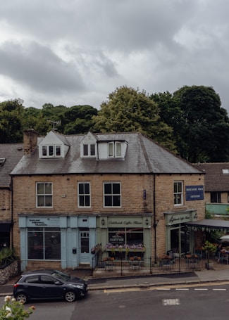 A two-story building made of stone bricks, featuring small businesses on the ground floor including a personal training center, a coffee shop with outdoor seating, and an interior design store. The sky is overcast with clouds, and there is a parked car on the street in front of the building. Lush green trees are visible in the background.