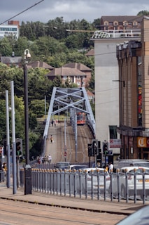A city street scene featuring a tram crossing a bridge with people walking alongside. The area is surrounded by greenery and urban buildings, including an apartment complex and a commercial building with a 'To Let' sign. Traffic lights and vehicles are visible, indicating a busy urban environment.