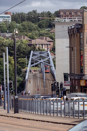 A city street scene featuring a tram crossing a bridge with people walking alongside. The area is surrounded by greenery and urban buildings, including an apartment complex and a commercial building with a 'To Let' sign. Traffic lights and vehicles are visible, indicating a busy urban environment.