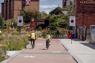 Cyclists wearing reflective vests ride down a shared pedestrian and bike path lined with greenery. Buildings with signage can be seen in the background, and there are restricted parking zone signs on both sides of the path. People are visible walking in the background, and the area is surrounded by urban architecture with some graffiti on the walls.