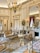 A lavishly decorated room featuring ornate furnishings, gilded accents, and intricate patterned rugs. A grand chandelier hangs from the ceiling, while oil portraits, mirrors, and a marble fireplace enhance the elegant atmosphere. Richly upholstered chairs, small tables, and decorative statues add to the luxurious ambiance.