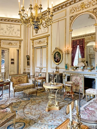 A lavishly decorated room featuring ornate furnishings, gilded accents, and intricate patterned rugs. A grand chandelier hangs from the ceiling, while oil portraits, mirrors, and a marble fireplace enhance the elegant atmosphere. Richly upholstered chairs, small tables, and decorative statues add to the luxurious ambiance.