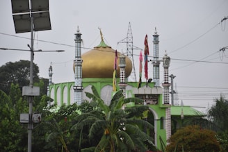 A mosque with a prominent golden dome and four tall minarets decorated with colorful flags stands amid lush green vegetation. Utility poles and power lines are visible in the background, contrasting with the religious architecture.