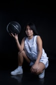 A person wearing a basketball uniform squats while holding a black basketball in one hand. The uniform features the number 23 and a recognizable team name. The setting is dimly lit against a dark backdrop, highlighting the subject.