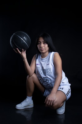 A person wearing a basketball uniform squats while holding a black basketball in one hand. The uniform features the number 23 and a recognizable team name. The setting is dimly lit against a dark backdrop, highlighting the subject.