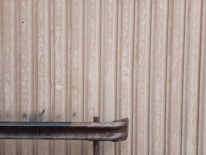 A metal wall with a weathered and rusted pipe or railing in front of it. The wall has vertical corrugated lines with a worn-out appearance, featuring faded paint and some dark stains.