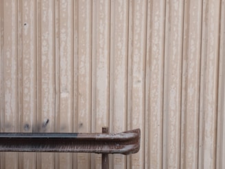 A metal wall with a weathered and rusted pipe or railing in front of it. The wall has vertical corrugated lines with a worn-out appearance, featuring faded paint and some dark stains.