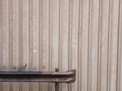 A metal wall with a weathered and rusted pipe or railing in front of it. The wall has vertical corrugated lines with a worn-out appearance, featuring faded paint and some dark stains.