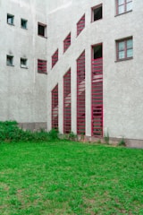 A building facade features a series of vertically aligned red-framed windows with unique angular designs. The walls of the structure are concrete with a textured finish. In front of the building, there is a small patch of green grass with some overgrown shrubs near the base of the building.