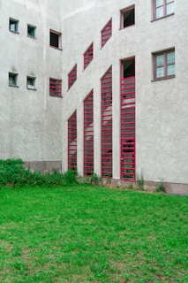A building facade features a series of vertically aligned red-framed windows with unique angular designs. The walls of the structure are concrete with a textured finish. In front of the building, there is a small patch of green grass with some overgrown shrubs near the base of the building.