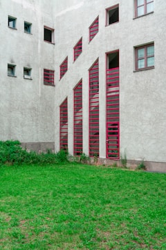A building facade features a series of vertically aligned red-framed windows with unique angular designs. The walls of the structure are concrete with a textured finish. In front of the building, there is a small patch of green grass with some overgrown shrubs near the base of the building.