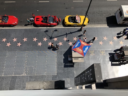 An aerial view captures the bustling sidewalk of the Hollywood Walk of Fame, displaying its iconic pink star tiles embedded into the pavement. A bright red sports car and a yellow convertible are parked along the street, with a group of people gathered near them. Pedestrians can be seen walking along the sidewalk, and a colorful tent provides shade for a stand displaying promotional materials. In the corner, a sign indicates the location of the Dolby Theatre.
