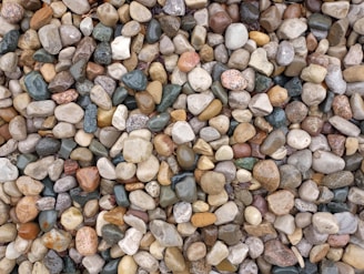A collection of variously colored pebbles and stones piled together. The stones range in size and shape, with smooth surfaces and natural textures. Colors include shades of gray, brown, beige, white, and hints of green.