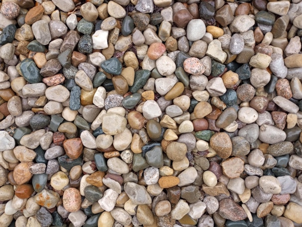 A collection of variously colored pebbles and stones piled together. The stones range in size and shape, with smooth surfaces and natural textures. Colors include shades of gray, brown, beige, white, and hints of green.