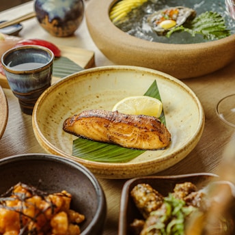 A beautifully arranged Japanese meal featuring a grilled salmon fillet garnished with a lemon slice on a plate with a leaf. Surrounding dishes include a bowl of ice with an oyster and garnish, a cup of sake, a dish of mixed appetizers, and sushi pieces. The setting conveys a luxurious and appetizing dining experience.