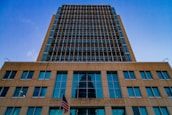 A tall modern building with a grid-like facade dominates the scene. The structure is made of glass and steel, reflecting the blue sky above. The lower section of the building bears the inscription 'United States of America' with an American flag displayed prominently in front.