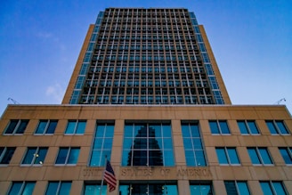 A tall modern building with a grid-like facade dominates the scene. The structure is made of glass and steel, reflecting the blue sky above. The lower section of the building bears the inscription 'United States of America' with an American flag displayed prominently in front.