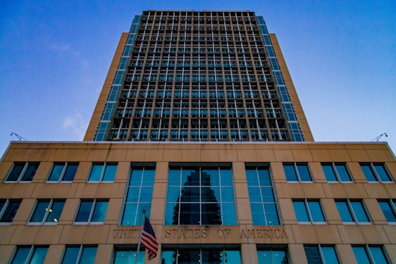 A tall modern building with a grid-like facade dominates the scene. The structure is made of glass and steel, reflecting the blue sky above. The lower section of the building bears the inscription 'United States of America' with an American flag displayed prominently in front.