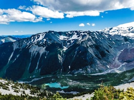 A breathtaking mountainous landscape featuring snow-capped peaks under a partly cloudy blue sky. The rugged terrain is complemented by a serene turquoise lake nestled among pine forests. The scene captures the natural beauty and grandeur of a high-altitude environment.