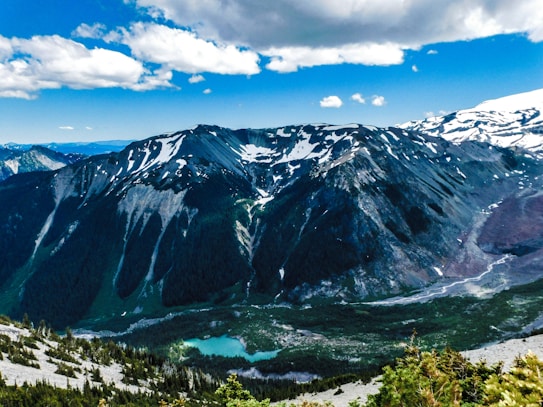 A breathtaking mountainous landscape featuring snow-capped peaks under a partly cloudy blue sky. The rugged terrain is complemented by a serene turquoise lake nestled among pine forests. The scene captures the natural beauty and grandeur of a high-altitude environment.