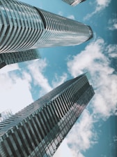 Tall, modern skyscrapers with sleek glass facades reach towards a bright blue sky filled with scattered fluffy clouds.