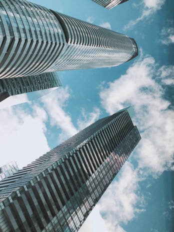 Tall, modern skyscrapers with sleek glass facades reach towards a bright blue sky filled with scattered fluffy clouds.
