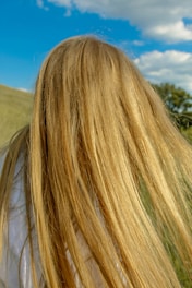 Long, flowing blonde hair is captured in close-up against the backdrop of a bright blue sky with scattered clouds. The scene appears to be set outdoors, possibly in a grassy field.