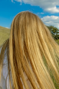Long, flowing blonde hair is captured in close-up against the backdrop of a bright blue sky with scattered clouds. The scene appears to be set outdoors, possibly in a grassy field.