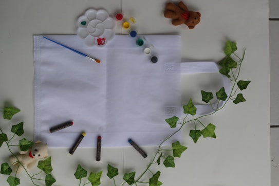 A flat white cloth bag is placed on a table surrounded by art supplies, including a paintbrush, a palette with various colors of paint, and several oil pastels. Two small plush toys, one bear and one dog, are positioned at opposite corners. Green ivy leaves are arranged decoratively around the bag.