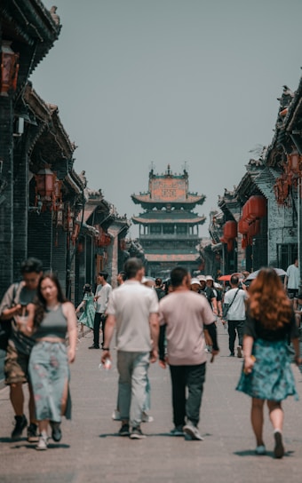 A street scene in an ancient town with traditional Chinese architecture, featuring a busy pedestrian path lined with historic buildings adorned with red lanterns. A group of people walks along the street, showcasing a blend of modern and traditional attire.