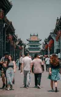 A street scene in an ancient town with traditional Chinese architecture, featuring a busy pedestrian path lined with historic buildings adorned with red lanterns. A group of people walks along the street, showcasing a blend of modern and traditional attire.
