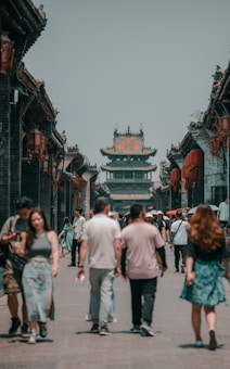 A street scene in an ancient town with traditional Chinese architecture, featuring a busy pedestrian path lined with historic buildings adorned with red lanterns. A group of people walks along the street, showcasing a blend of modern and traditional attire.
