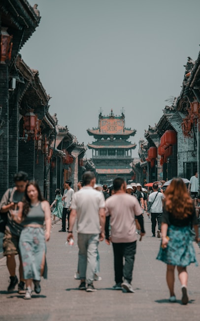 A street scene in an ancient town with traditional Chinese architecture, featuring a busy pedestrian path lined with historic buildings adorned with red lanterns. A group of people walks along the street, showcasing a blend of modern and traditional attire.