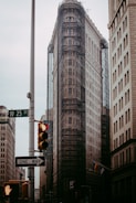 A tall, iconic triangular building is under renovation with scaffolding covering most of its surface. It is flanked by other older high-rise buildings in an urban setting. A traffic light with a red signal and a one-way street sign are visible in the foreground. An overcast sky adds to the mood of the scene.