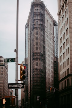 A tall, iconic triangular building is under renovation with scaffolding covering most of its surface. It is flanked by other older high-rise buildings in an urban setting. A traffic light with a red signal and a one-way street sign are visible in the foreground. An overcast sky adds to the mood of the scene.