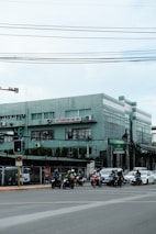 A street scene featuring a group of motorcyclists and a car waiting at a traffic light at an intersection. A green building with the sign 'EASTERNSTAR LINE, INC.' is visible in the background. Overhead power lines stretch across the top of the image. The area appears urban with additional signage and various structures in sight.