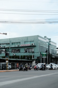 A street scene featuring a group of motorcyclists and a car waiting at a traffic light at an intersection. A green building with the sign 'EASTERNSTAR LINE, INC.' is visible in the background. Overhead power lines stretch across the top of the image. The area appears urban with additional signage and various structures in sight.