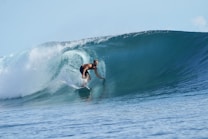 A surfer skillfully rides inside a large, powerful ocean wave. The wave curls above the surfer, creating a natural tunnel. The water is clear and blue, evoking a sense of dynamic motion and energy.