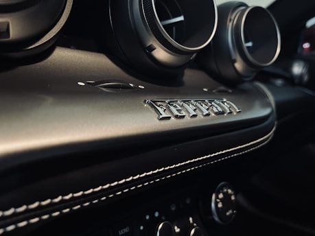 A close-up view of a car's interior, featuring air vents and detailed stitching on the dashboard. Prominently displayed is an emblem spelling 'Ferrari', conveying luxury and attention to detail.