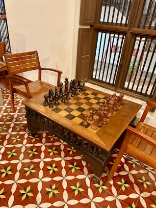A wooden chessboard set is positioned on a matching wooden table, surrounded by wooden chairs with a classic design. The floor features a decorative, geometric tile pattern with red, white, and black colors. Nearby, a window with vertical bars allows a view of the outside.