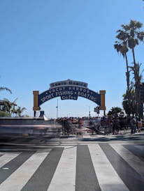 A bustling scene at the entrance of Santa Monica Pier, featuring a prominent arched sign that reads 'Santa Monica Yacht Harbor Sport Fishing Boating Cafes'. A crosswalk with blurred motion captures the movement of people, suggesting a busy atmosphere. Tall palm trees frame the scene, and the bright, clear sky adds to a lively Southern California vibe.