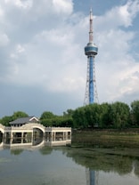 A tall broadcasting tower stands under a partially cloudy sky, with a traditional bridge and a body of water in the foreground. Surrounding the bridge are lush green trees, creating a pleasing contrast with the blue and gray tones of the tower and sky.