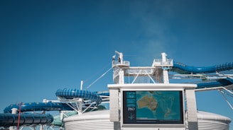 A large waterslide complex with blue and white tubing structure set against a clear blue sky. In the foreground, a digital map display shows a map of Australia and nearby regions, with a logo on the bottom left. A person is seen on the elevated walkway, highlighting the scale of the water slides.