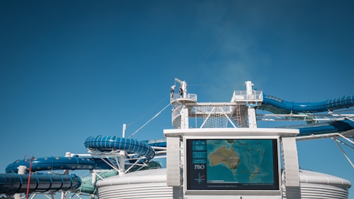 A large waterslide complex with blue and white tubing structure set against a clear blue sky. In the foreground, a digital map display shows a map of Australia and nearby regions, with a logo on the bottom left. A person is seen on the elevated walkway, highlighting the scale of the water slides.