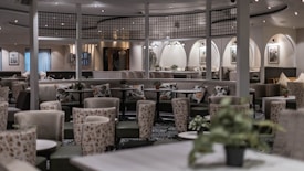 An elegant and modern restaurant or lounge area with a blend of contemporary and classic design elements. The room features multiple round tables surrounded by plush chairs with floral patterns, and soft lighting which creates a cozy ambiance. Decorative plants are placed on tables and around the room, contributing to a fresh and inviting atmosphere. The walls are adorned with framed artwork and soft, neutral colors dominate the decor. The ceiling has overhead lights arranged in a stylish manner, complementing the sophisticated setting.