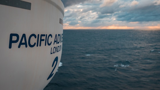 A life buoy with the text 'PACIFIC ADVENTURE LONDON 2' is prominently visible against a backdrop of an expansive ocean and a sky filled with clouds. The horizon separates the deep blue sea from the hazy sky featuring hints of warm colors near the horizon, suggesting either sunrise or sunset.