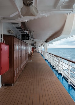 A long wooden deck on a ship featuring wooden lockers on the left, outdoor seating further along, and metal railings on the right overlooking the sea. The sky is partly cloudy, and the sea appears calm.