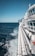 A large cruise ship is navigating through the ocean with its deck and railings prominently visible. The ship's white structure contrasts with the deep blue of the sea. Onboard amenities like a waterslide are visible in the background. The sky is clear and bright, indicating pleasant weather.
